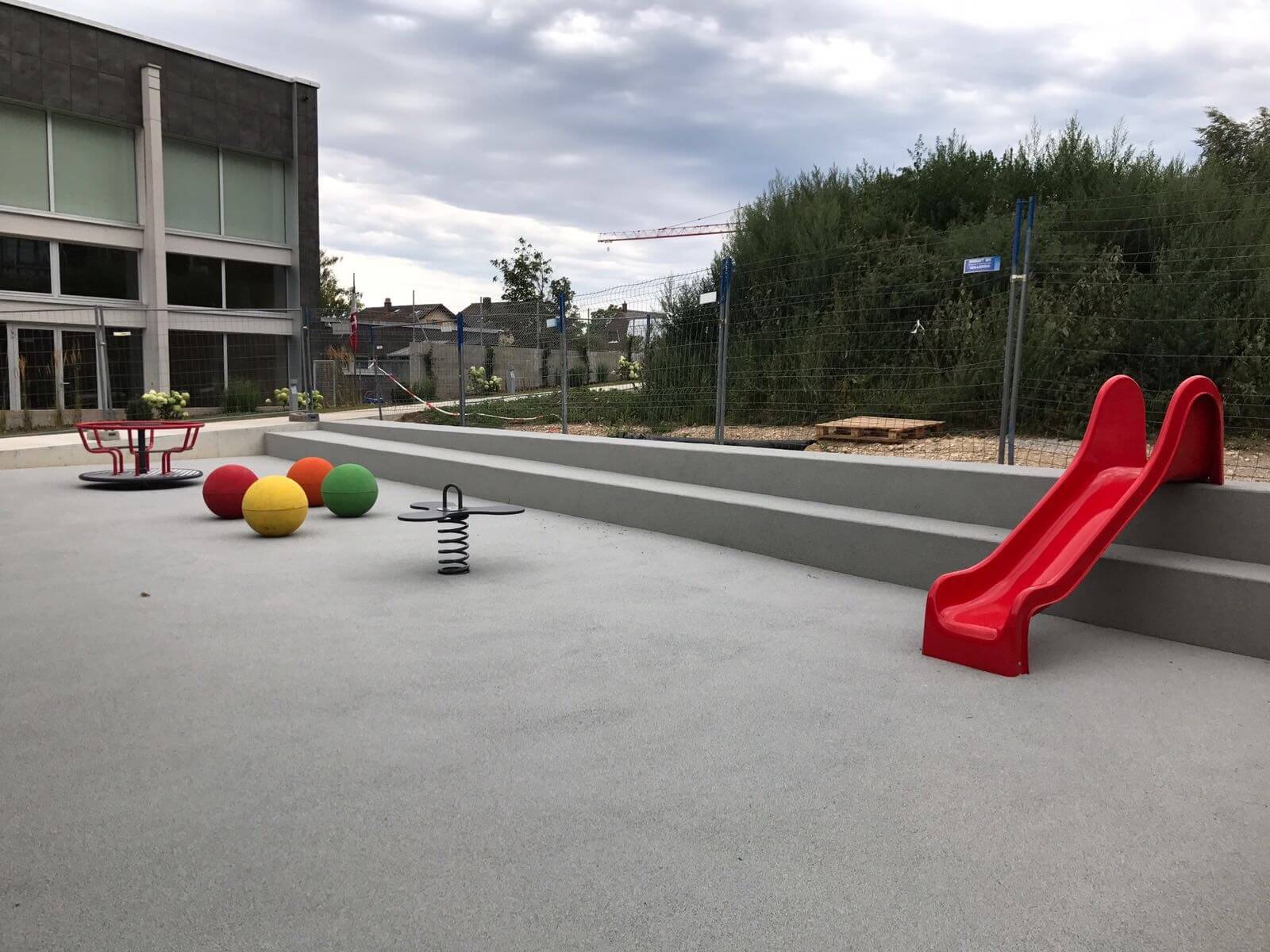 Ein moderner Spielplatz mit einer roten Rutsche, bunten Bällen, einem Karussell und einer Wippe auf einem sicheren Bodenbelag. Ideal für Kinder zum Spielen im Freien.