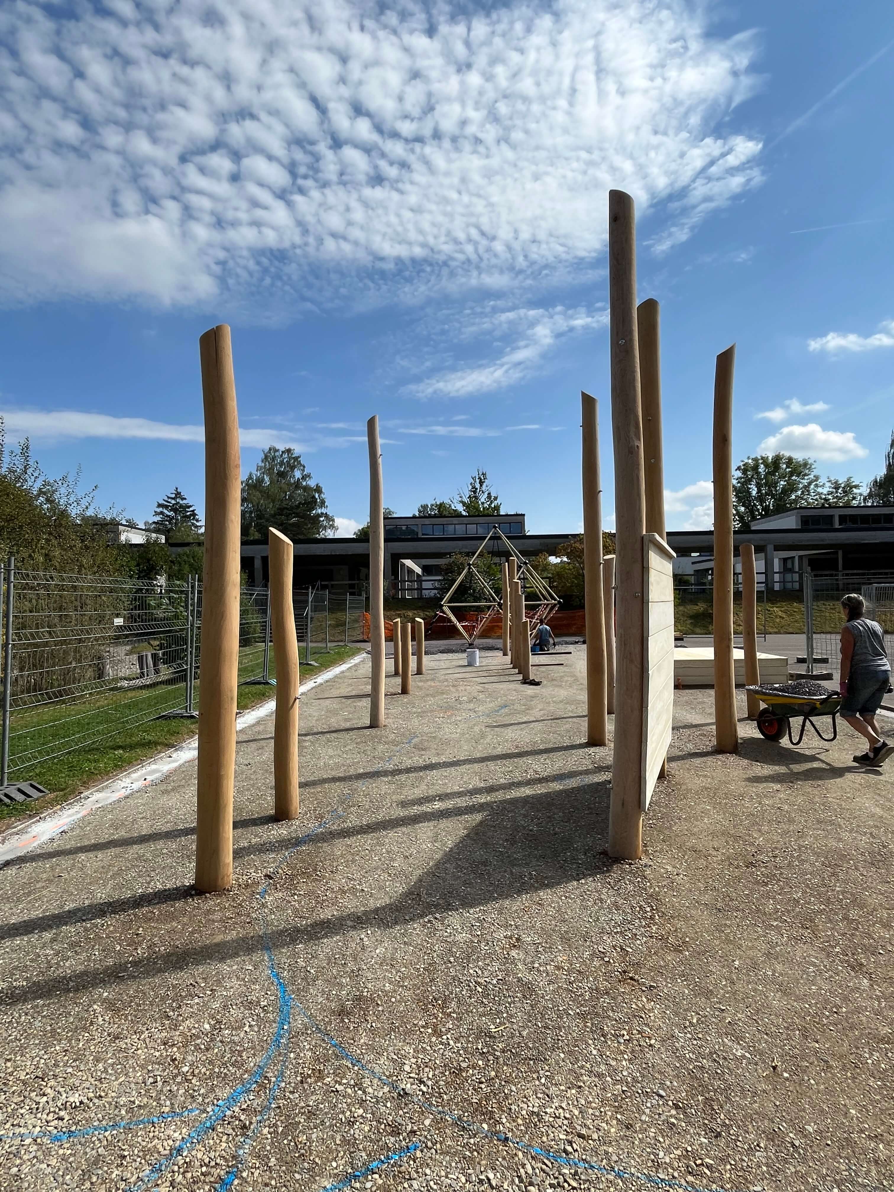 Bau eines Spielplatzes mit großen Holzstrukturen und einem Arbeiter, der eine Schubkarre schiebt. Der Himmel ist blau mit Wolken, und es ist ein sonniger Tag.