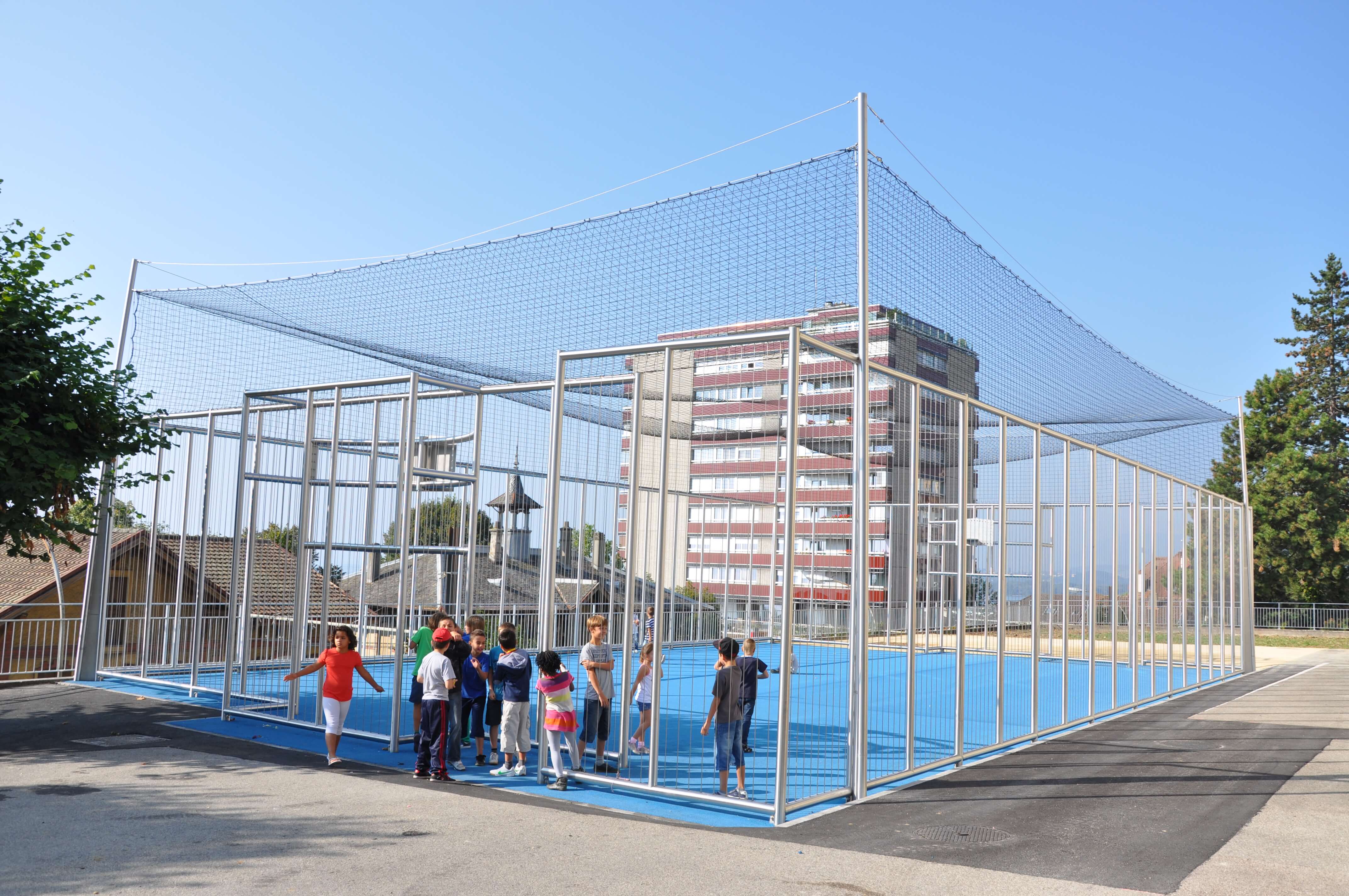 Gruppe von Kindern spielt auf einem umzäunten blauen Sportplatz im Freien, umgeben von Gebäuden
