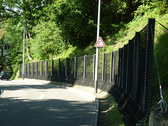 Eine Straße mit einer Kurve, die von einem hohen schwarzen Schutzzaun gesäumt ist, umgeben von üppigem Grün und einem Verkehrsschild.