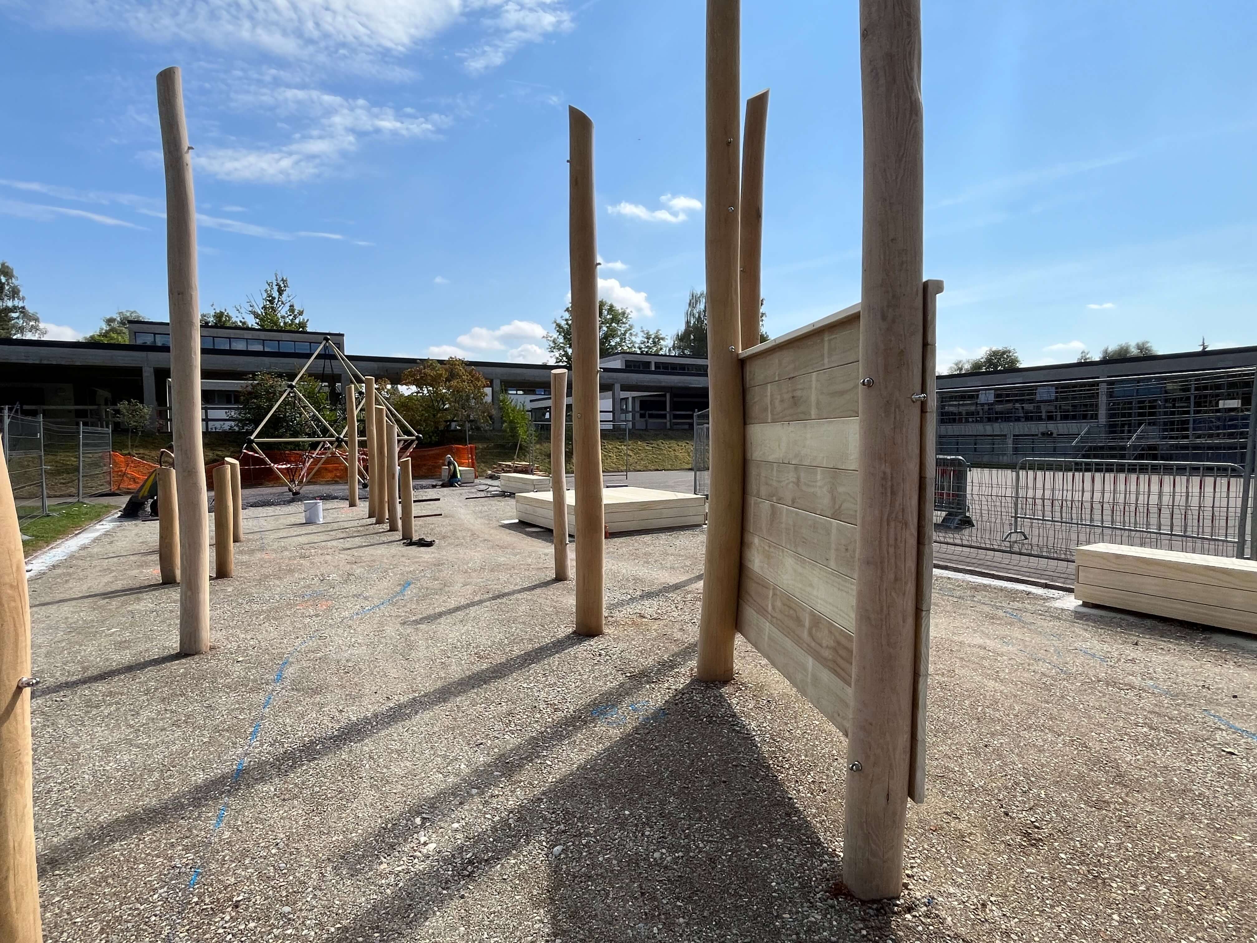 Ansicht eines im Bau befindlichen Spielplatzes mit mehreren aufrechten Holzpfosten und einem Klettergerüst im Hintergrund.