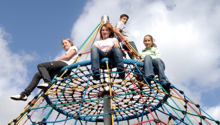 Vier Kinder klettern auf einem bunten, pyramidenförmigen Spielgerüst aus Seilen vor einem wolkigen Himmel