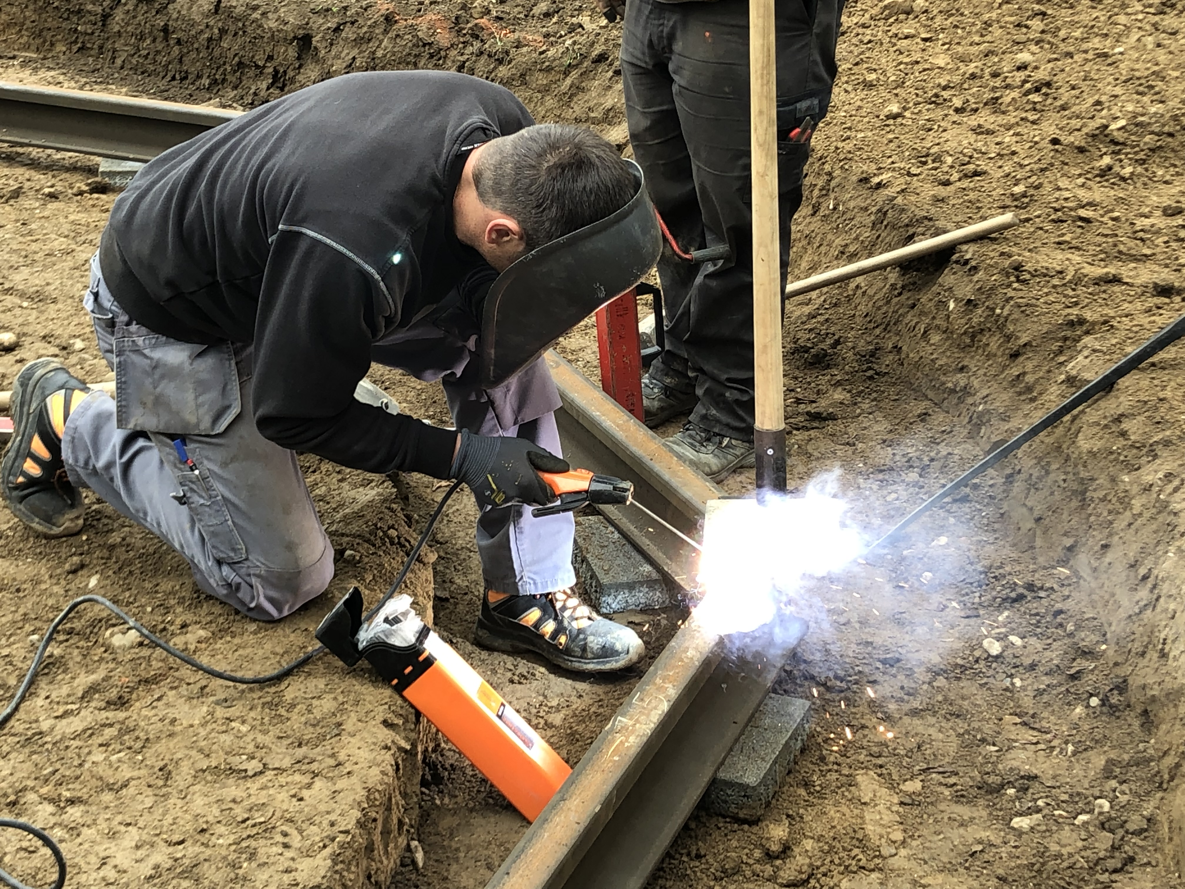 Ein Arbeiter mit Schutzmaske schweißt eine Schiene auf einer Baustelle. Funken fliegen, während er kniet und konzentriert arbeitet.