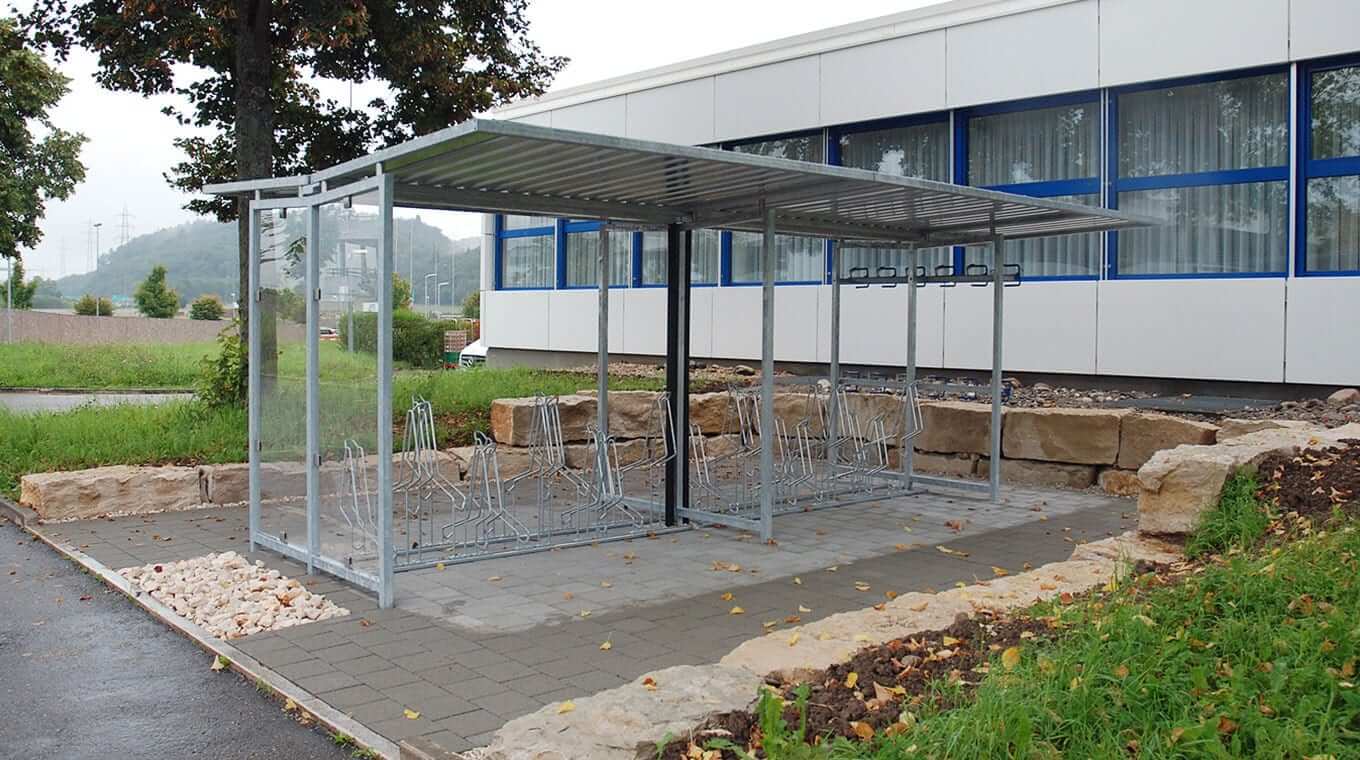 Ein moderner Fahrradunterstand aus Metall mit Überdachung, gelegen neben einem Bürogebäude, umgeben von Grünflächen.