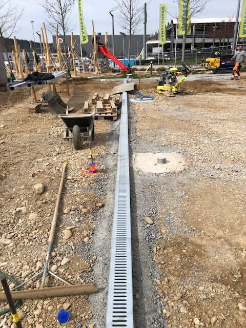 Chantier avec un caniveau de drainage installé, entouré de matériaux de construction et d'outils, avec une aire de jeux en cours de construction en arrière-plan