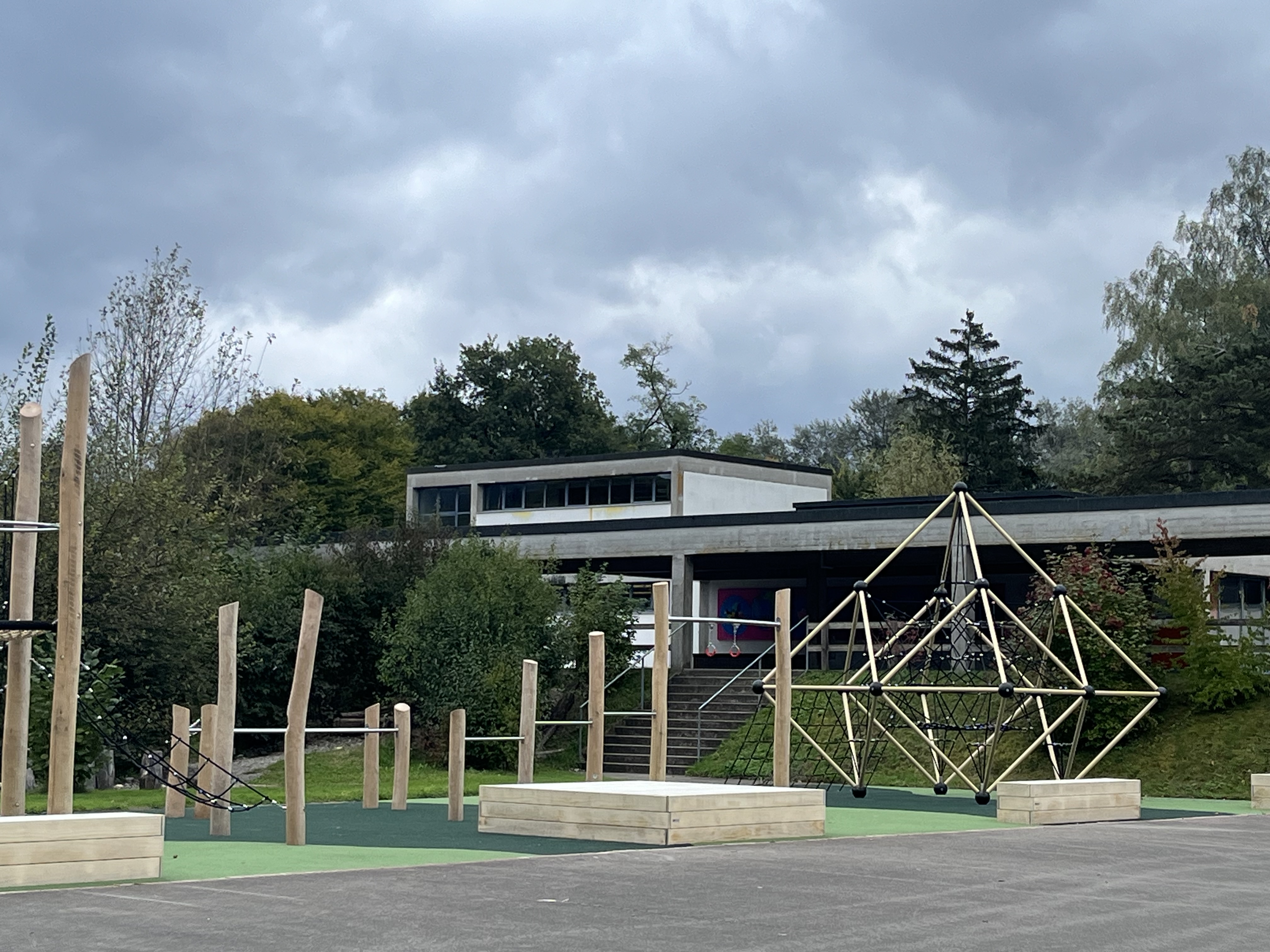 Ein moderner Spielplatz mit Holzklettergerüst und Seilen, gelegen vor einem flachen, modernen Gebäude unter bewölktem Himmel.