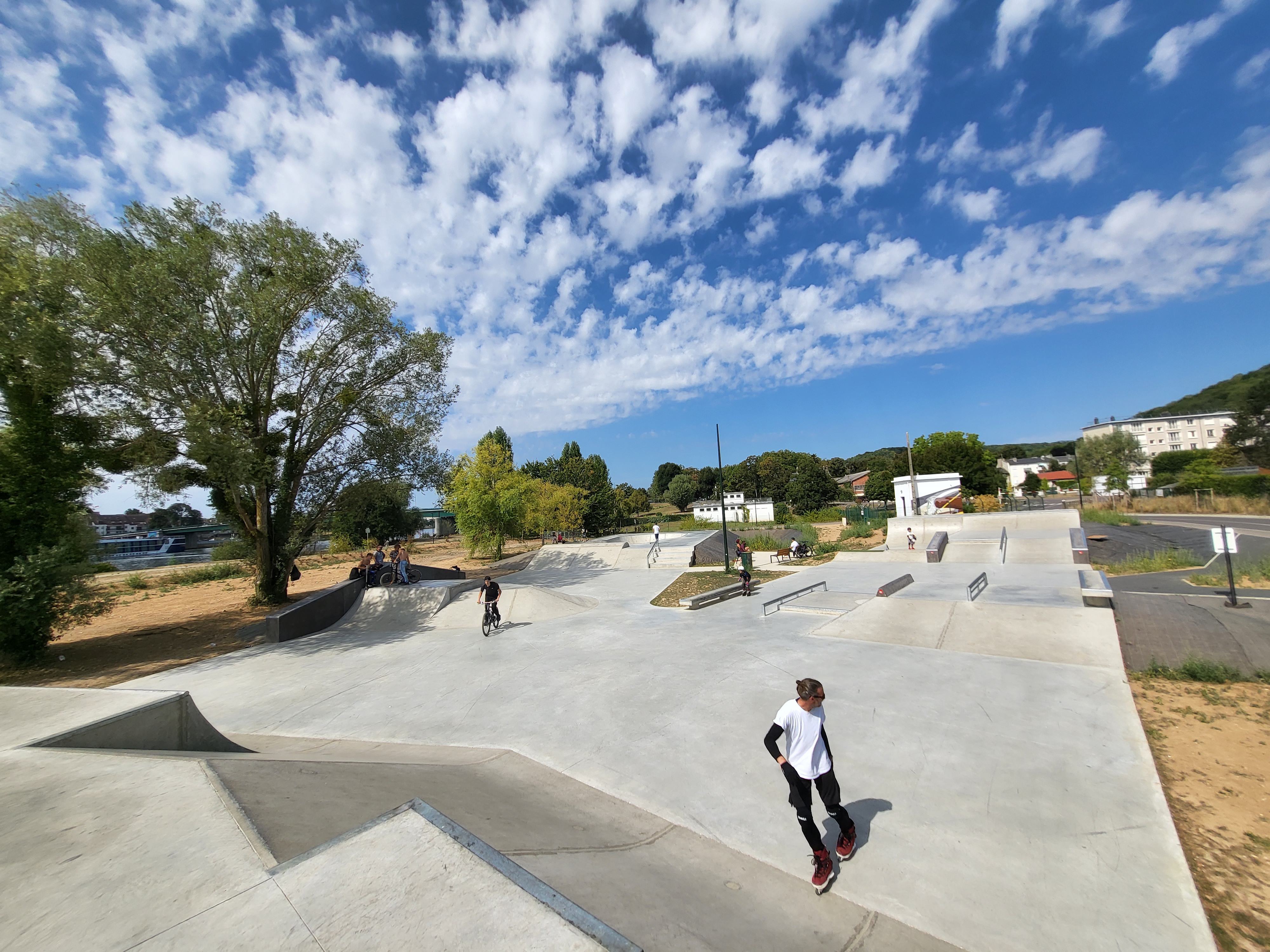 Un vaste skatepark avec des sportifs sous un ciel bleu nuageux, entouré d'arbres et de bâtiments.
