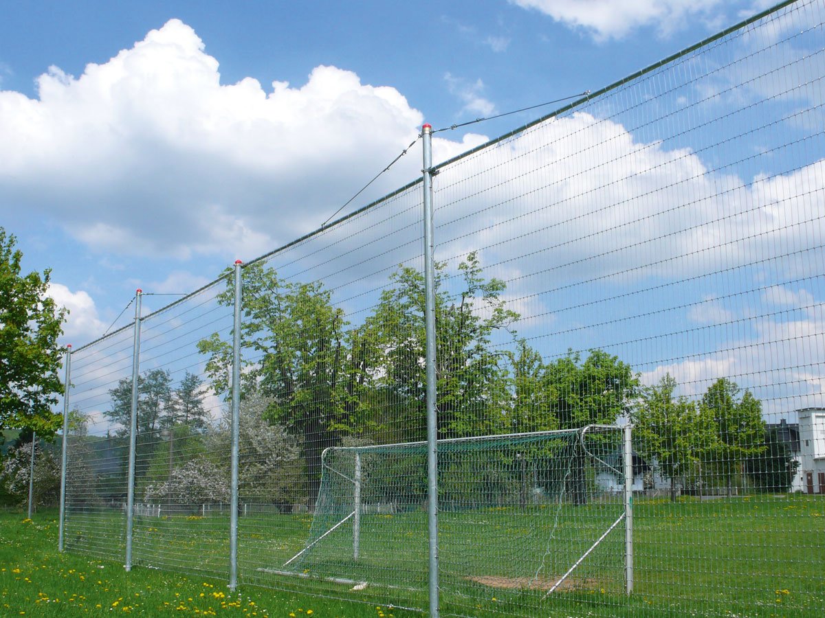 Hochsicherheitszaun auf einem Sportplatz mit grünem Rasen und blauem Himmel im Hintergrund