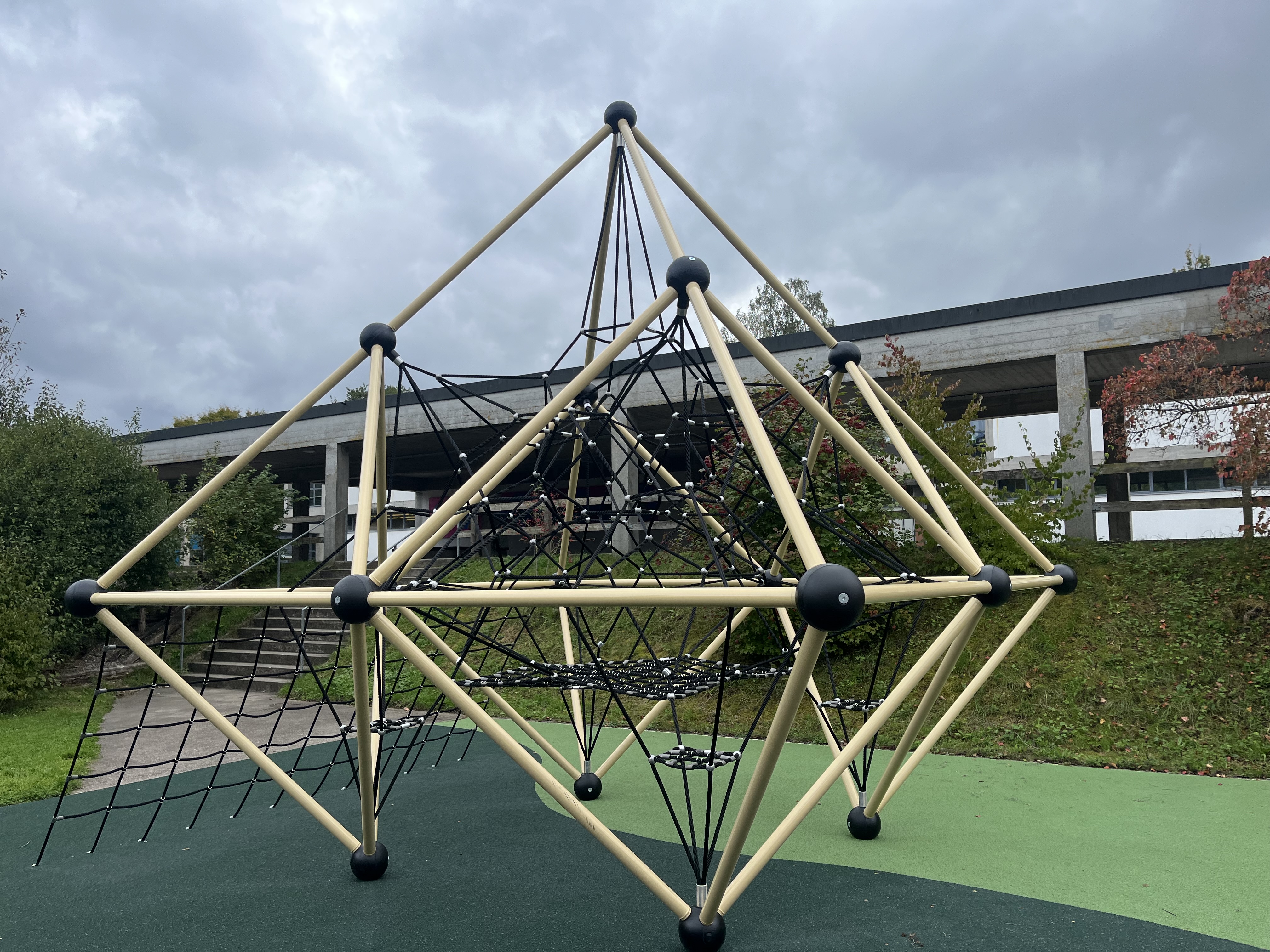 Ein modernes Klettergerüst aus Seilen und Stangen auf einem Spielplatz mit bewölktem Himmel im Hintergrund.