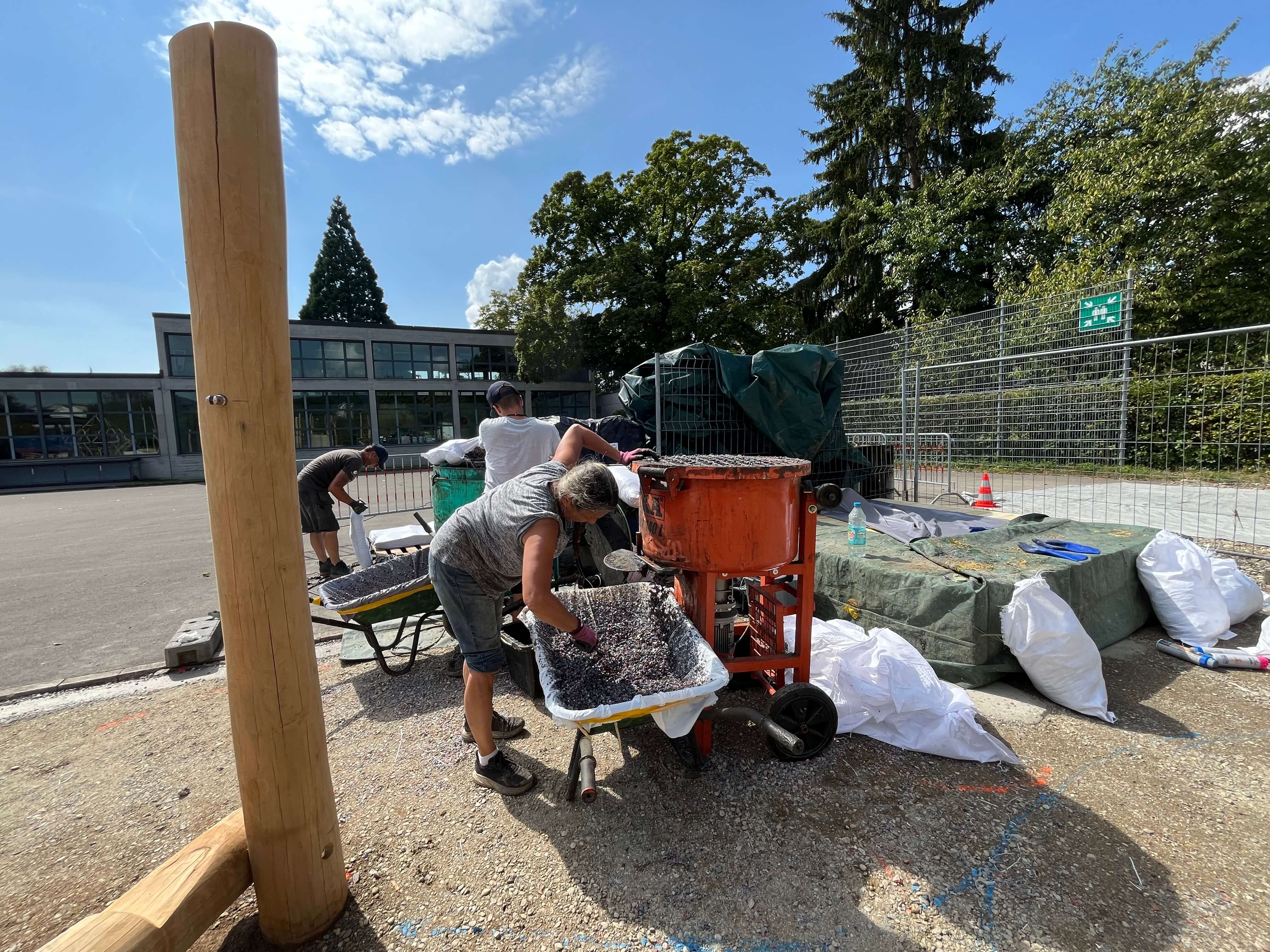 Personen mischen Baumaterialien mit einer Maschine auf einer Baustelle im Freien, umgeben von Baumaterialien und einem Gebäude im Hintergrund.