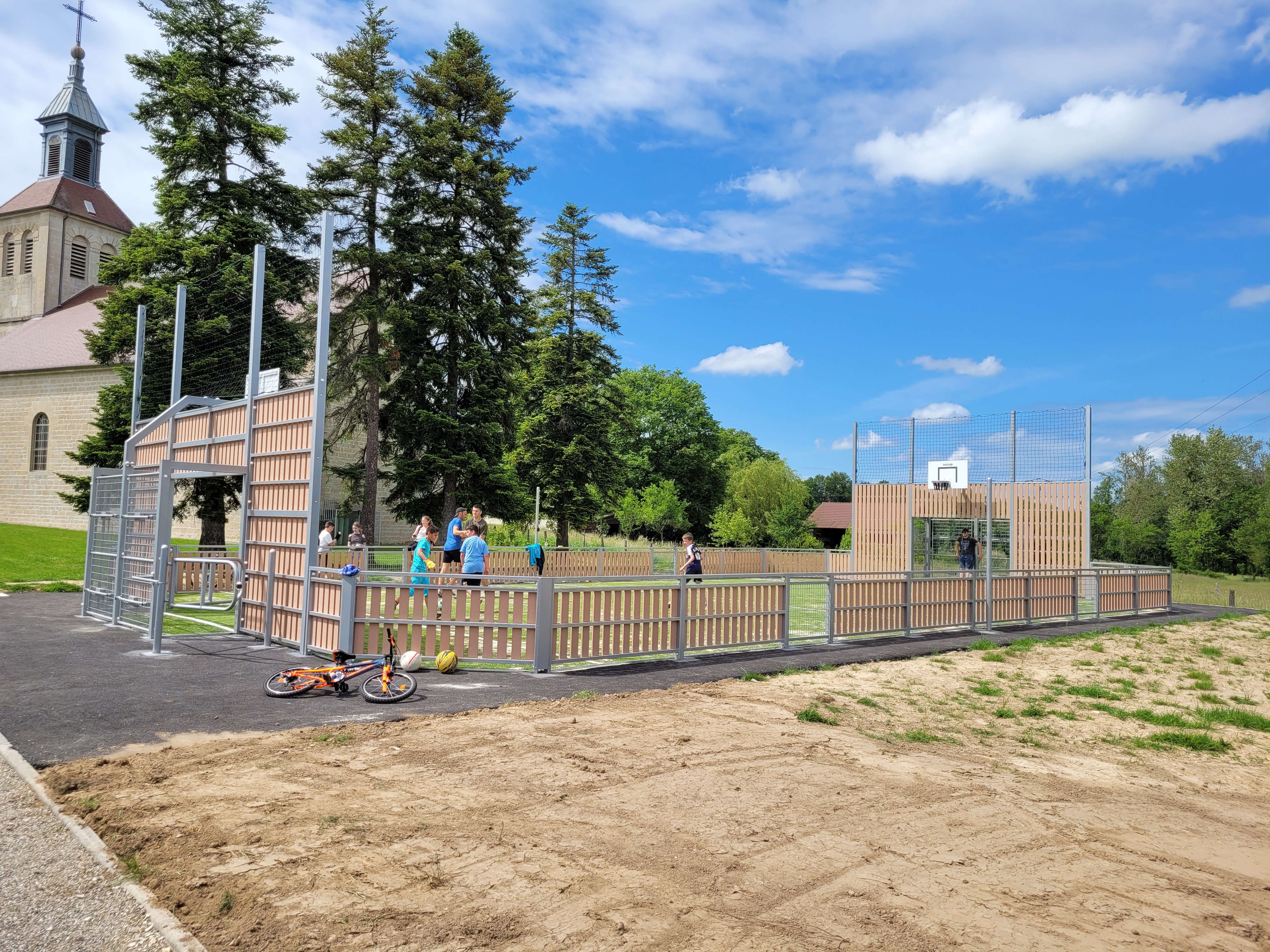 Plusieurs jeunes jouent sur un terrain de sport clôturé à côté d'une église, avec un vélo et des ballons au premier plan.