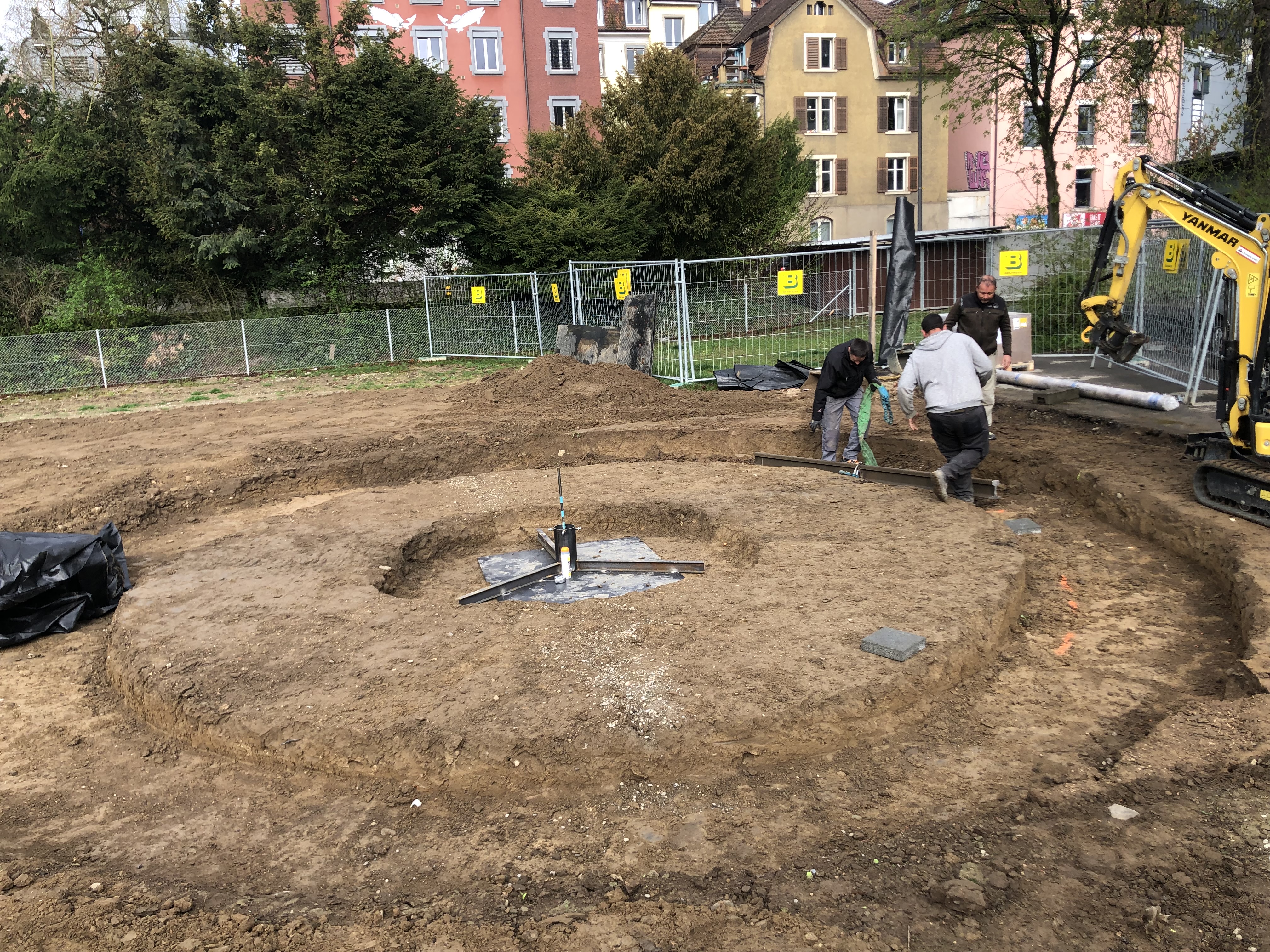 Drei Bauarbeiter arbeiten auf einer Baustelle an einem runden Fundament, umgeben von Erdreich und Bauzäunen.