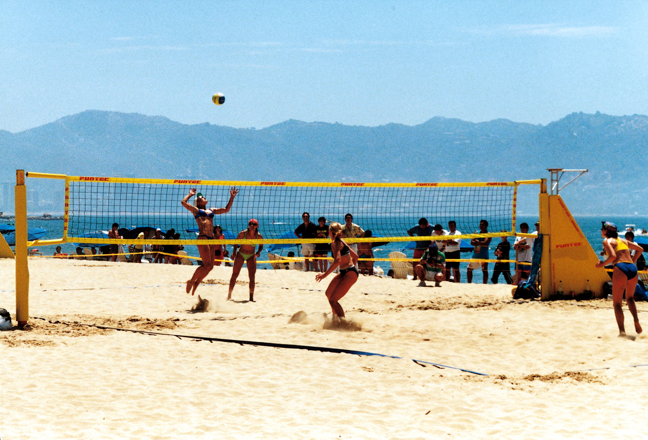 Quatre femmes disputent un match de beach-volley sur la plage, avec des spectateurs en arrière-plan, les montagnes et la mer au loin