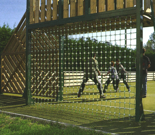 Eine Gruppe von Kindern spielt Fußball auf einem grünen Sportplatz, umgeben von einer Holzkonstruktion mit einem Netz.