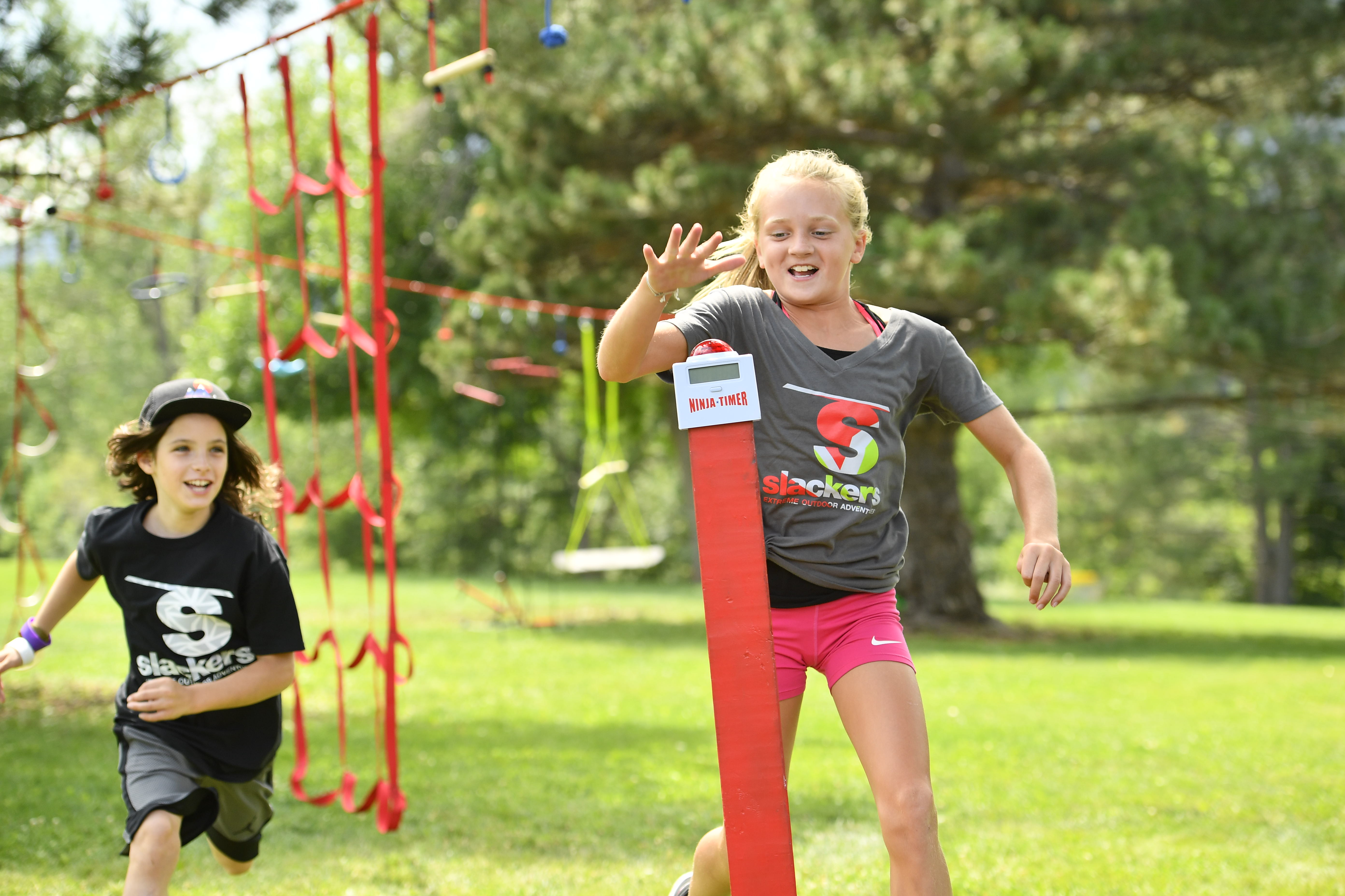 Zwei Kinder rennen fröhlich auf einem Outdoor-Spielplatz und erreichen einen Ninja-Timer-Pfosten.