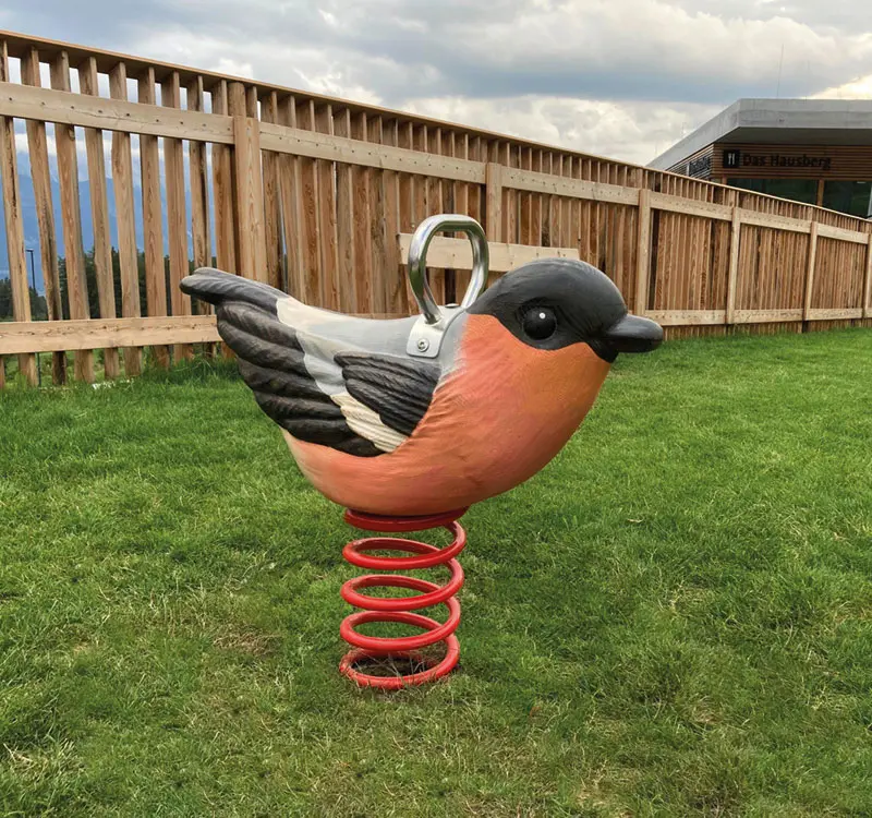 Ein Spielplatz-Wipptier in Form eines bunt bemalten Vogels auf einer Spiralfeder, platziert auf einer Rasenfläche vor einem Holzzaun.