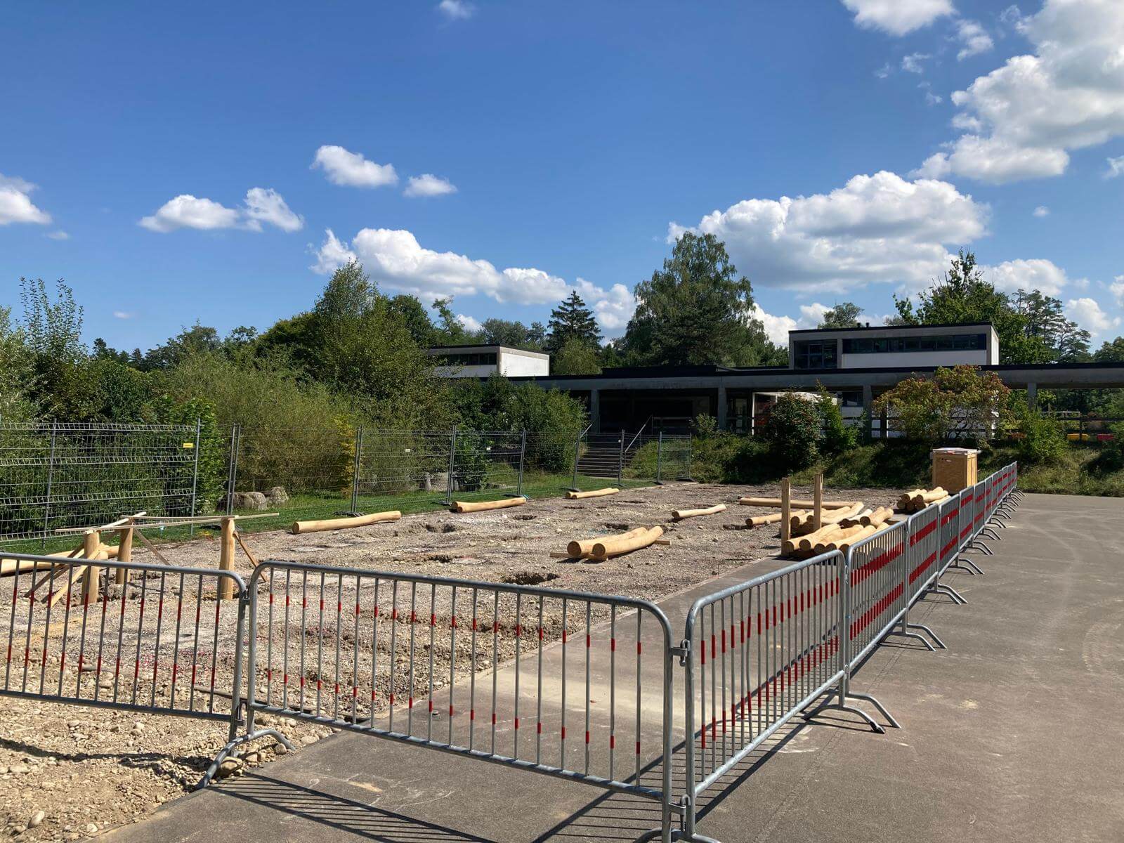Baustelle im Freien mit Metallzäunen und Holzstämmen, umgeben von Bäumen und einem Gebäude im Hintergrund