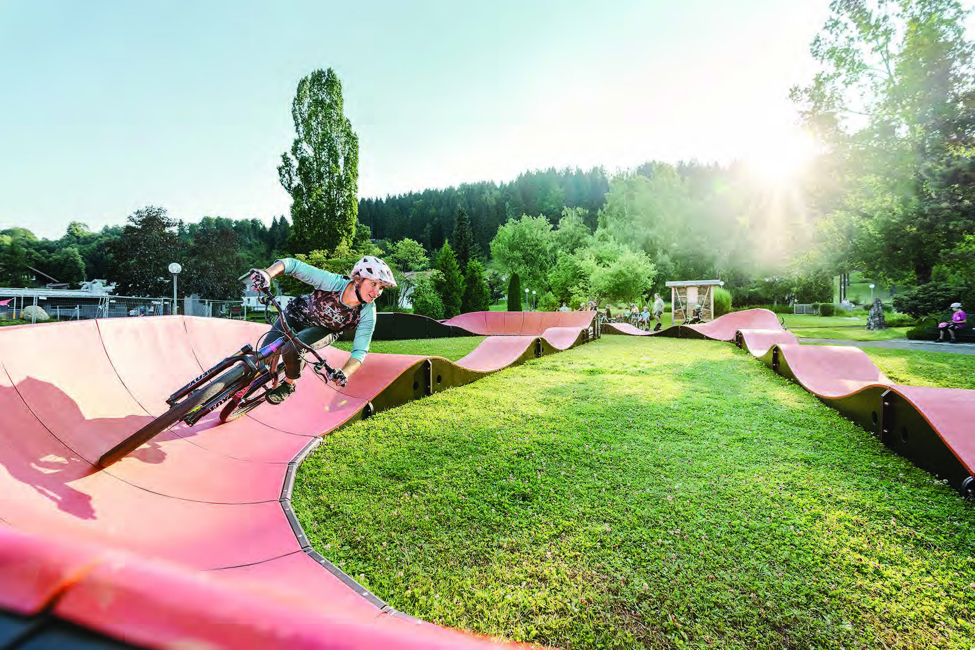 Un VTTiste casqué sur une pumptrack dans un paysage verdoyant, avec des arbres et des gens en arrière-plan