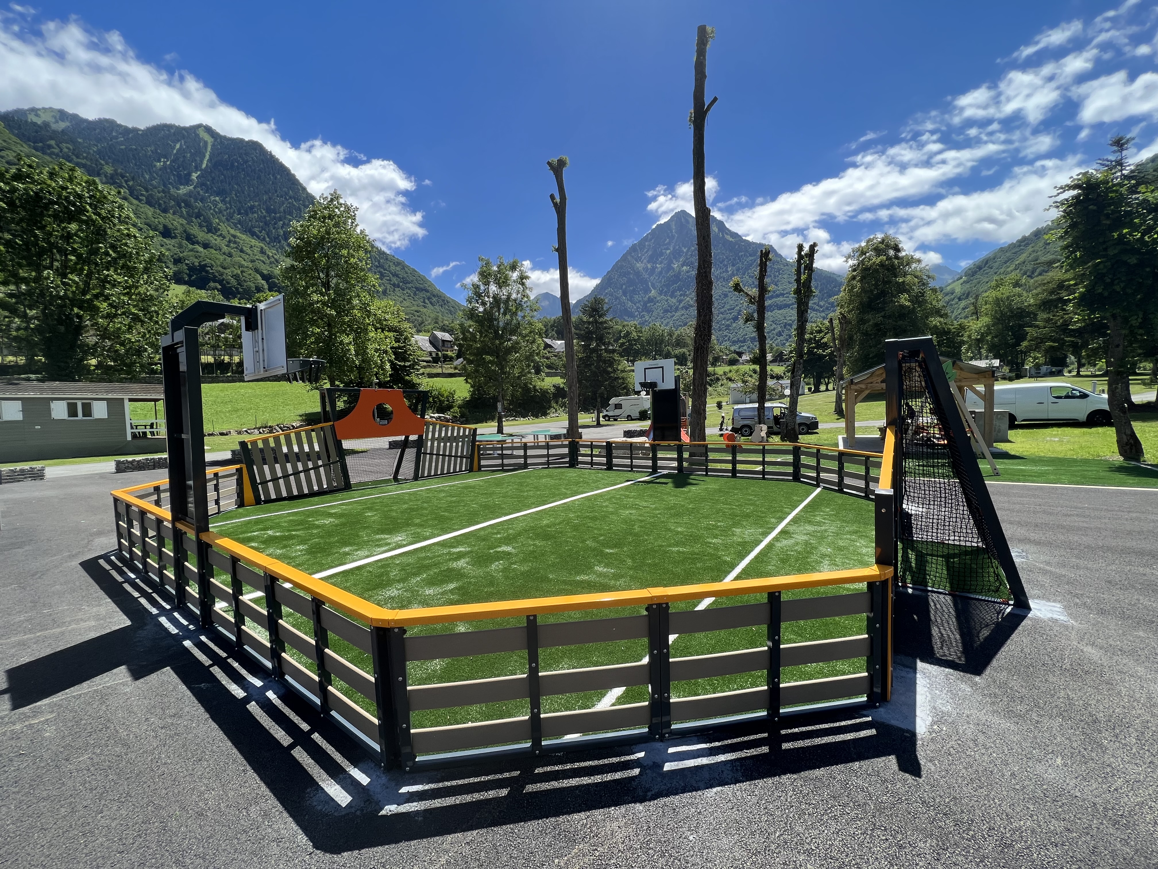 Un terrain de sport multifonctionnel moderne avec gazon synthétique et panier de basket, entouré d'un impressionnant paysage montagneux et d'un ciel bleu.