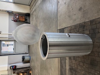Une grande poubelle cylindrique en acier inoxydable avec couvercle ouvert, posée dans un environnement d'atelier.