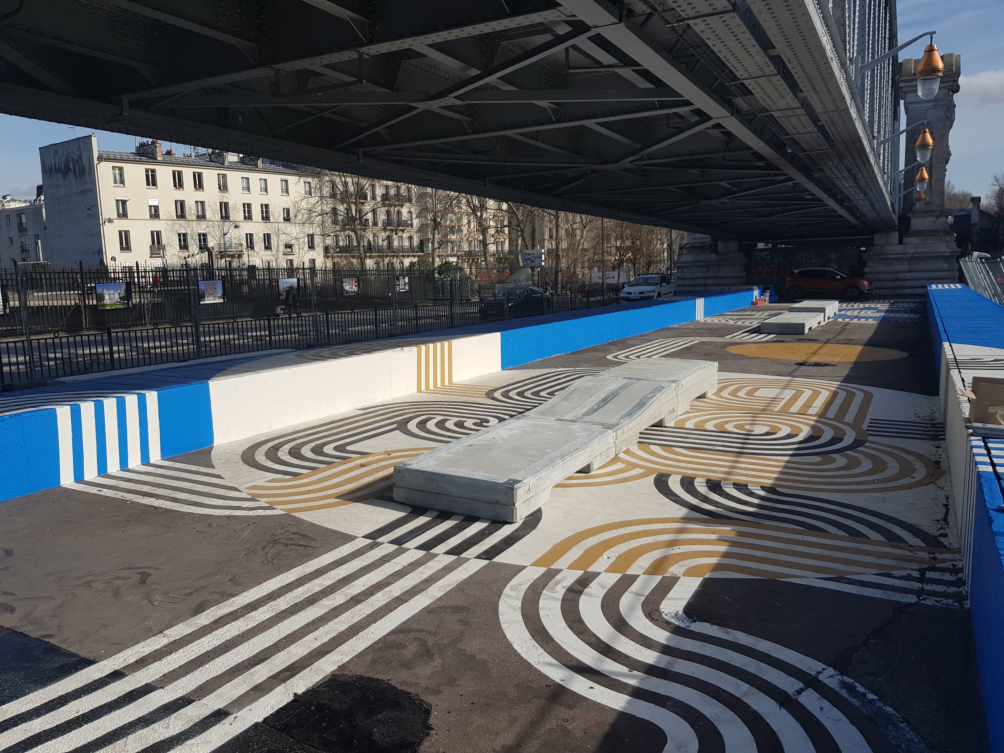 Peinture de rue artistique avec des motifs géométriques en noir, blanc et or sous un pont dans un environnement urbain.