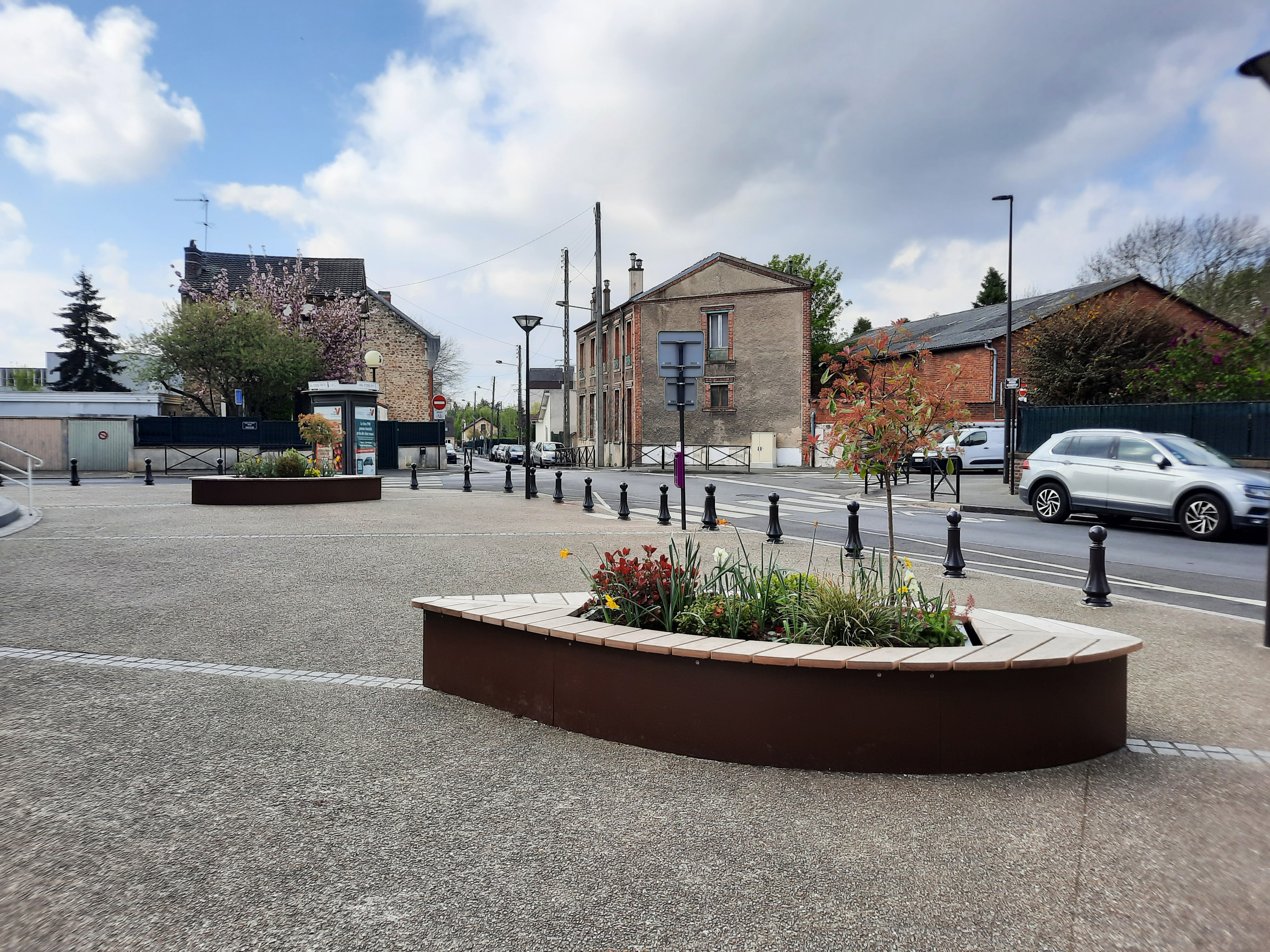 Leerer städtischer Platz mit Bänken und Blumenbeeten, umgeben von Wohngebäuden und einem vorbeifahrenden Auto
