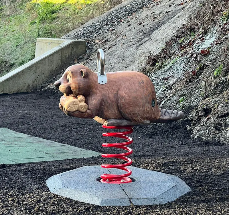 Eine Federwippe in Form eines Bären auf einem Spielplatz, montiert auf einer roten Feder und einem grauen Sockel, umgeben von Erde und Rasen.