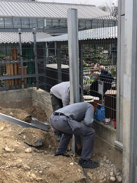 Deux ouvriers installent une clôture métallique sur un chantier, tandis qu'une exposition de plantes est visible en arrière-plan.