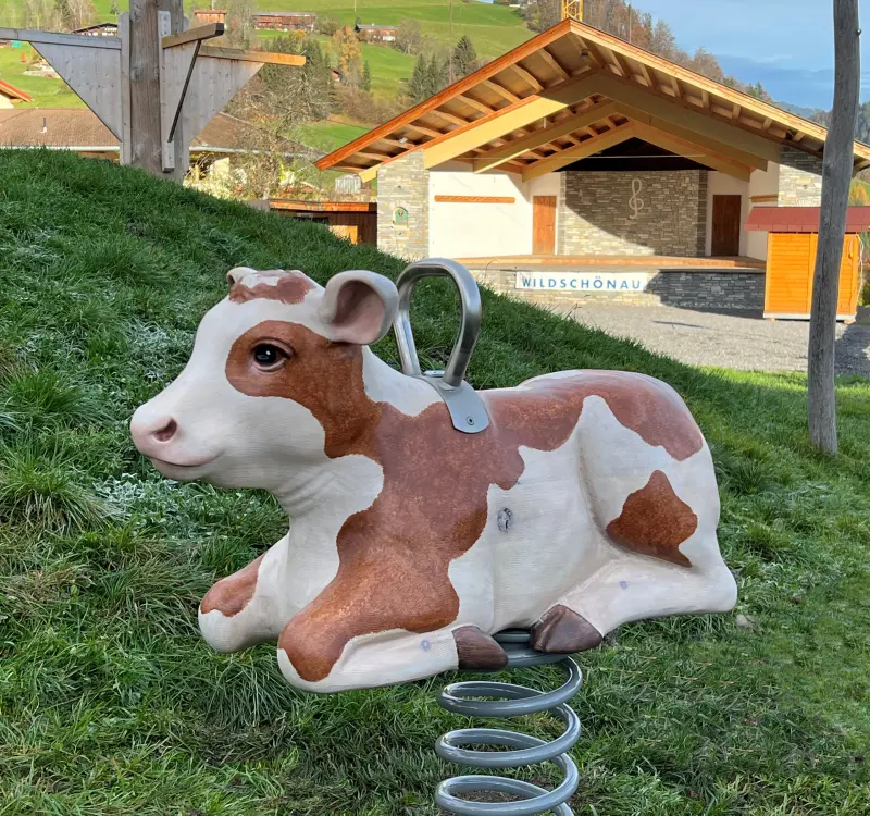 Eine Kuh-schaukel auf einem Spielplatz in Wildschönau, umgeben von grasbewachsenen Hügeln und einem Gebäude im Hintergrund.