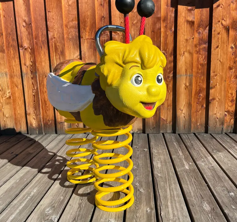 Gelbe Federwippe in Form einer Biene mit Flügeln und Fühlern, aufgestellt auf einem Holzdeck vor einer Holzwand.