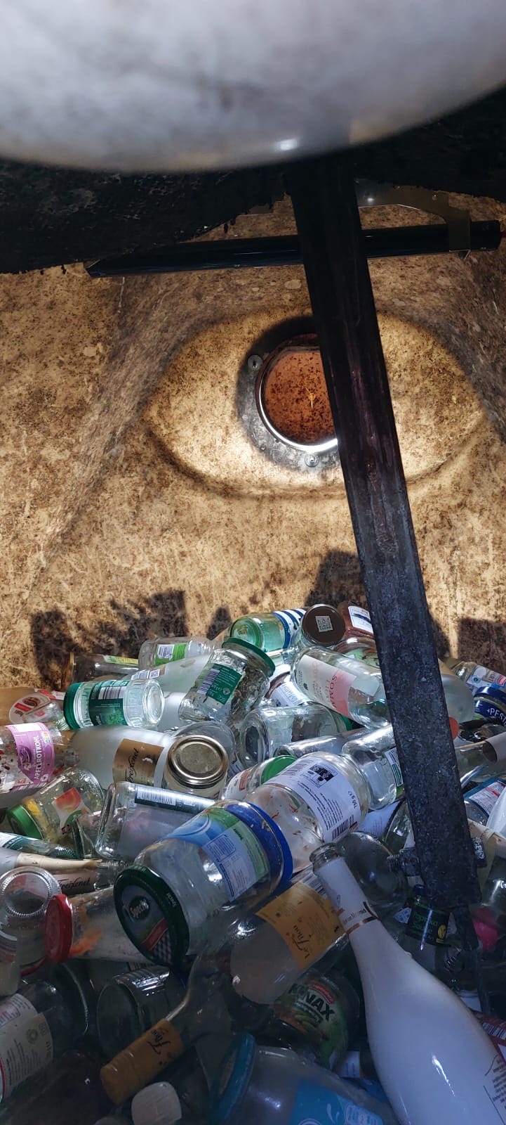 Verschiedene leere Glasflaschen und -gläser in einem Recyclingcontainer, teilweise Etiketten sichtbar, umgeben von einer runden Öffnung an der Oberseite.