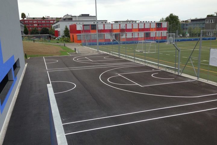 Une cour d'école moderne avec un terrain de basket-ball récemment aménagé et un terrain de football adjacent. Les bâtiments scolaires sont visibles en arrière-plan.