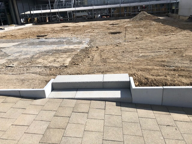 Eine Baustelle mit neu verlegten Bodenplatten und einer kleinen Treppe, die zu einem Erdreich führt. Im Hintergrund sind Baugerüste sichtbar.