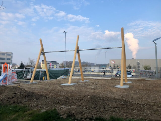 Eine Baustelle mit einem großen Holzgerüst auf einem unbebauten Gelände vor einem Industriegebäude.