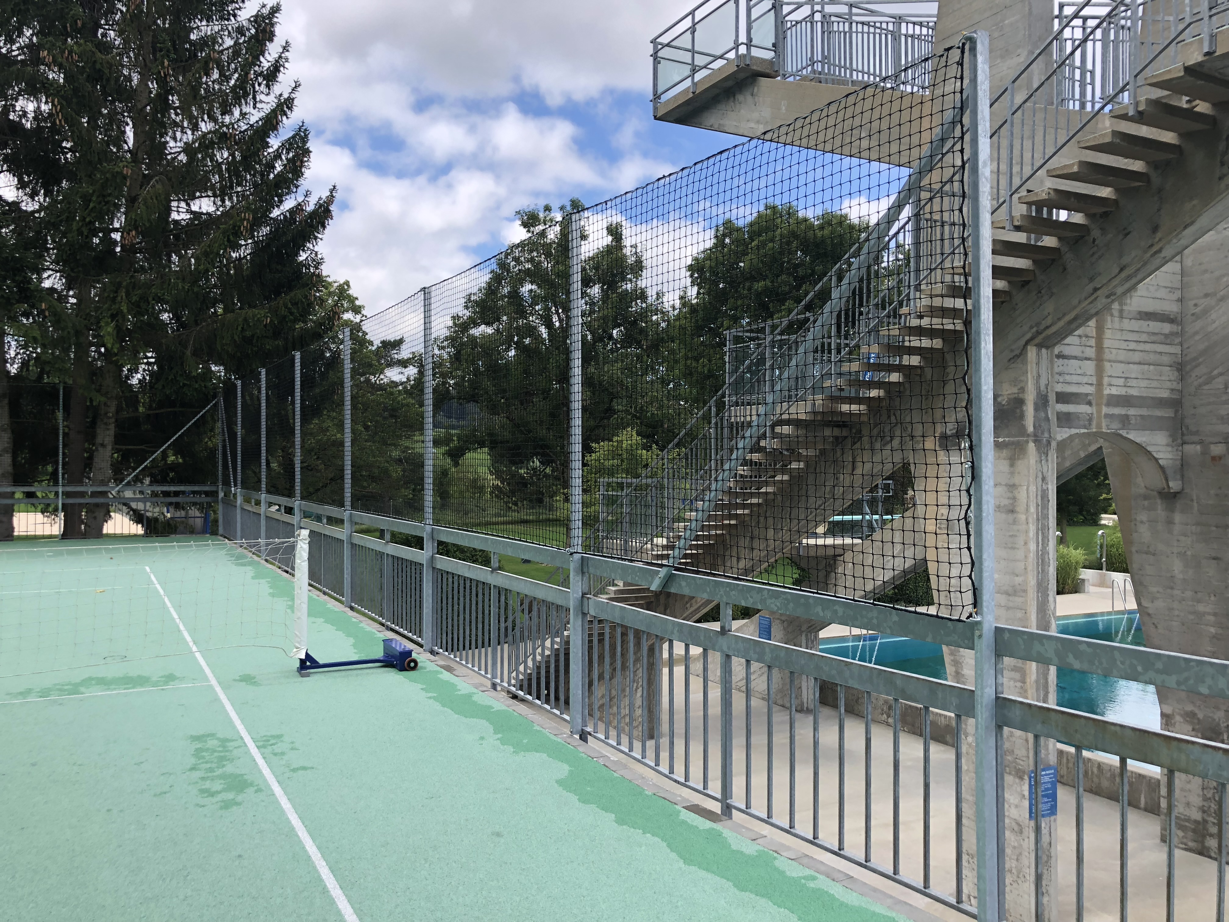 Ein leerer Tennisplatz mit grünem Boden und seitlichem Netz, umgeben von Bäumen und einer Beton-Treppe.