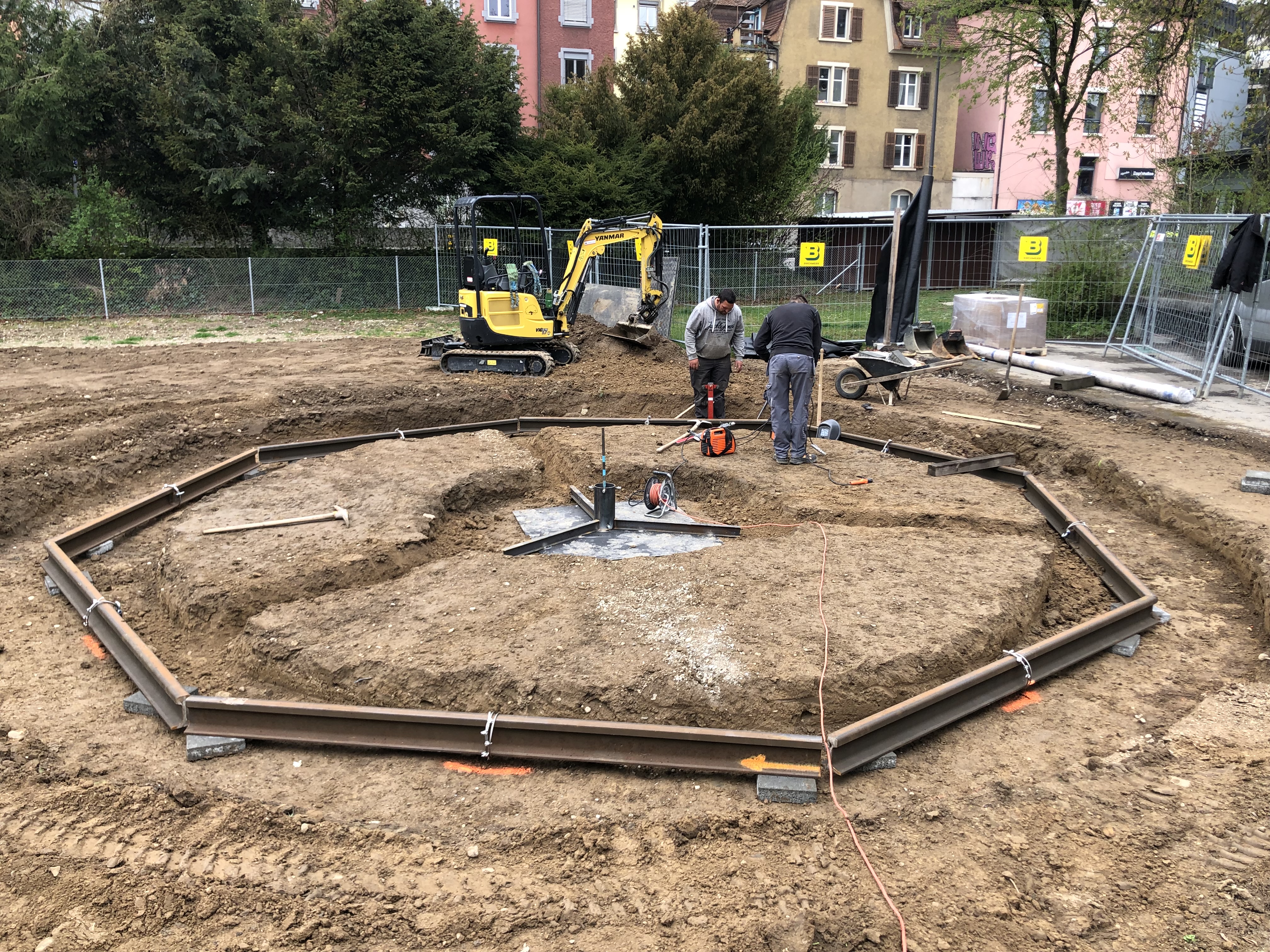 Zwei Bauarbeiter arbeiten an einer Baustelle im Freien, mit einem kleinen gelben Bagger und Metallrahmen im Vordergrund.