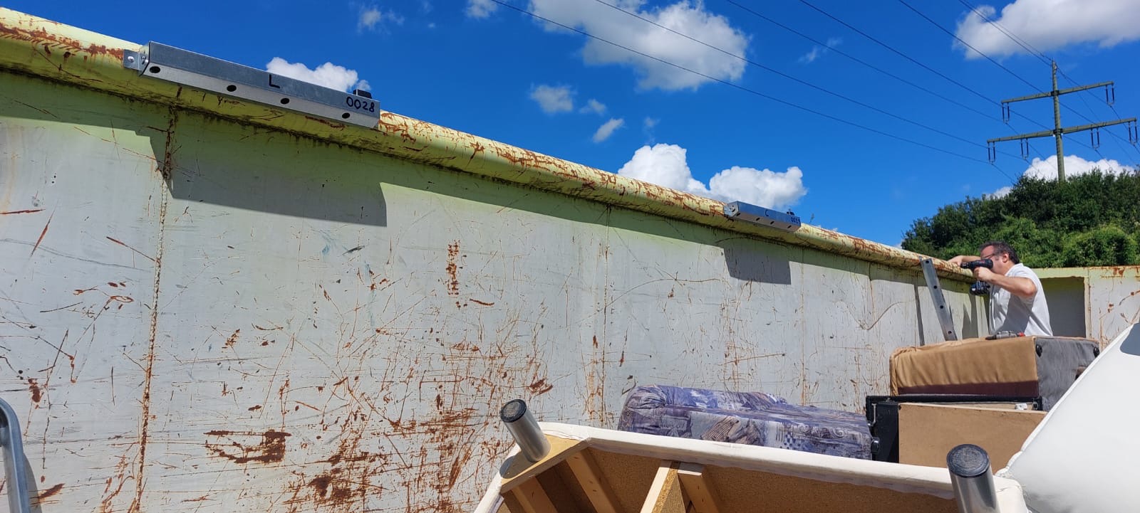 Ein Mann benutzt eine Bohrmaschine auf einer Leiter neben einem großen Container, Himmel mit Wolken im Hintergrund