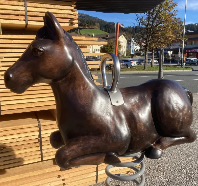 Ein aus Holz gefertigtes Pferd als Spielgerät auf einer Spiralfeder, platziert auf einem Freigelände vor einem Hintergrund mit Holzbrettern und Bäumen.