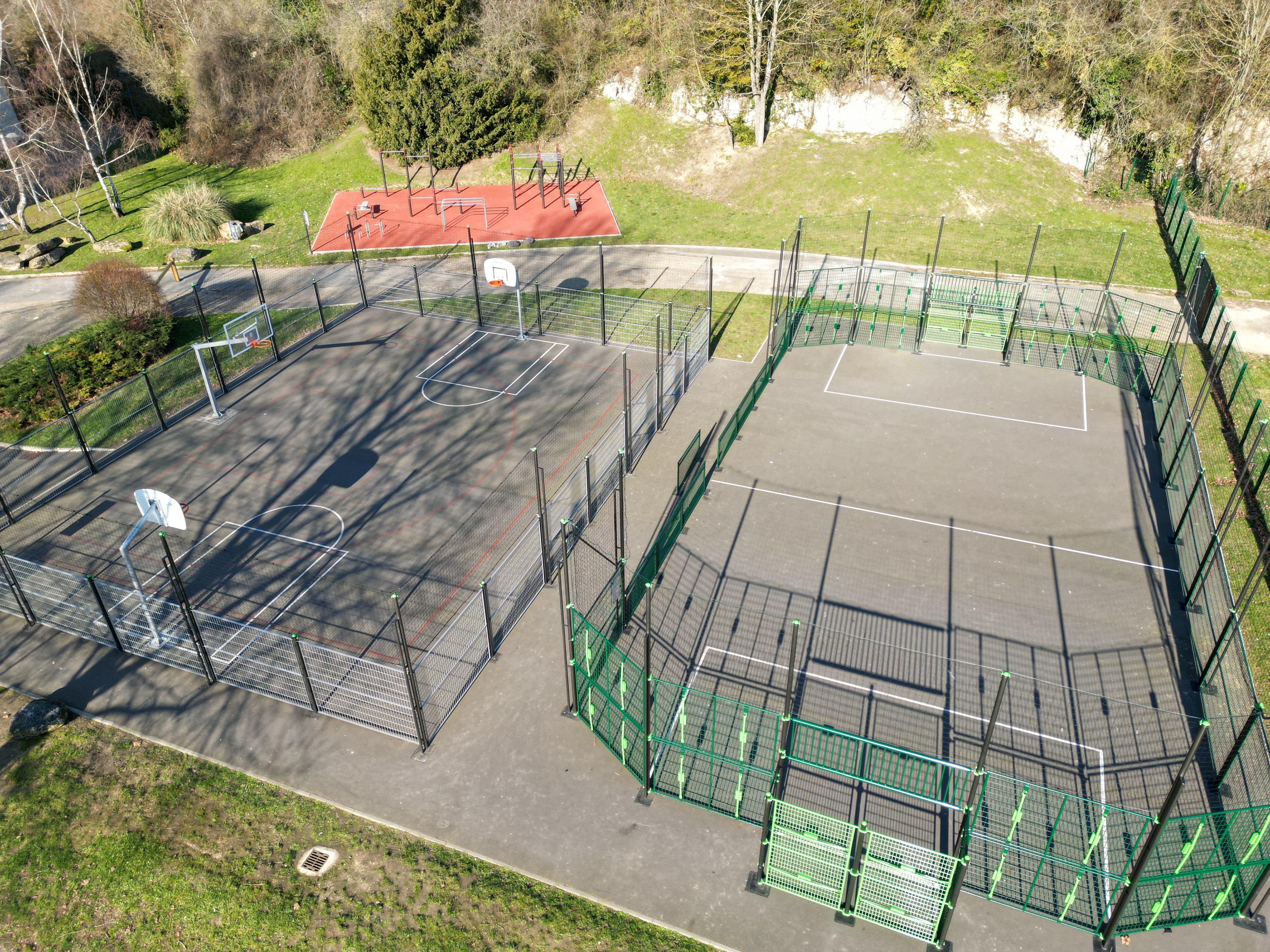 Vue aérienne d'un parc comprenant deux terrains de sport clôturés, un terrain de basket et un terrain polyvalent, entourés d'arbres et d'espaces verts.