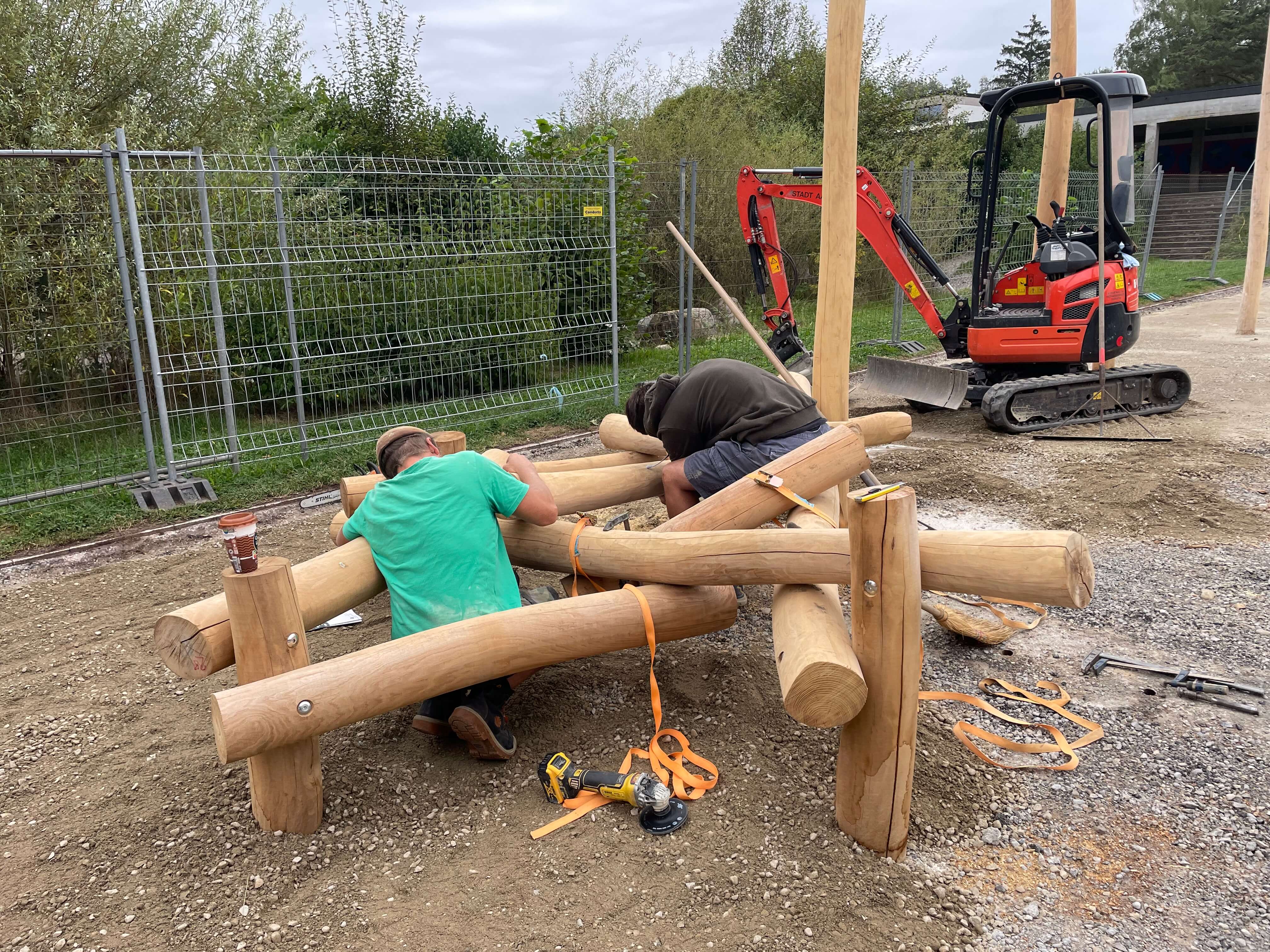 Zwei Personen arbeiten an der Montage von Holzpfosten auf einer Baustelle, ein kleiner Bagger steht daneben.