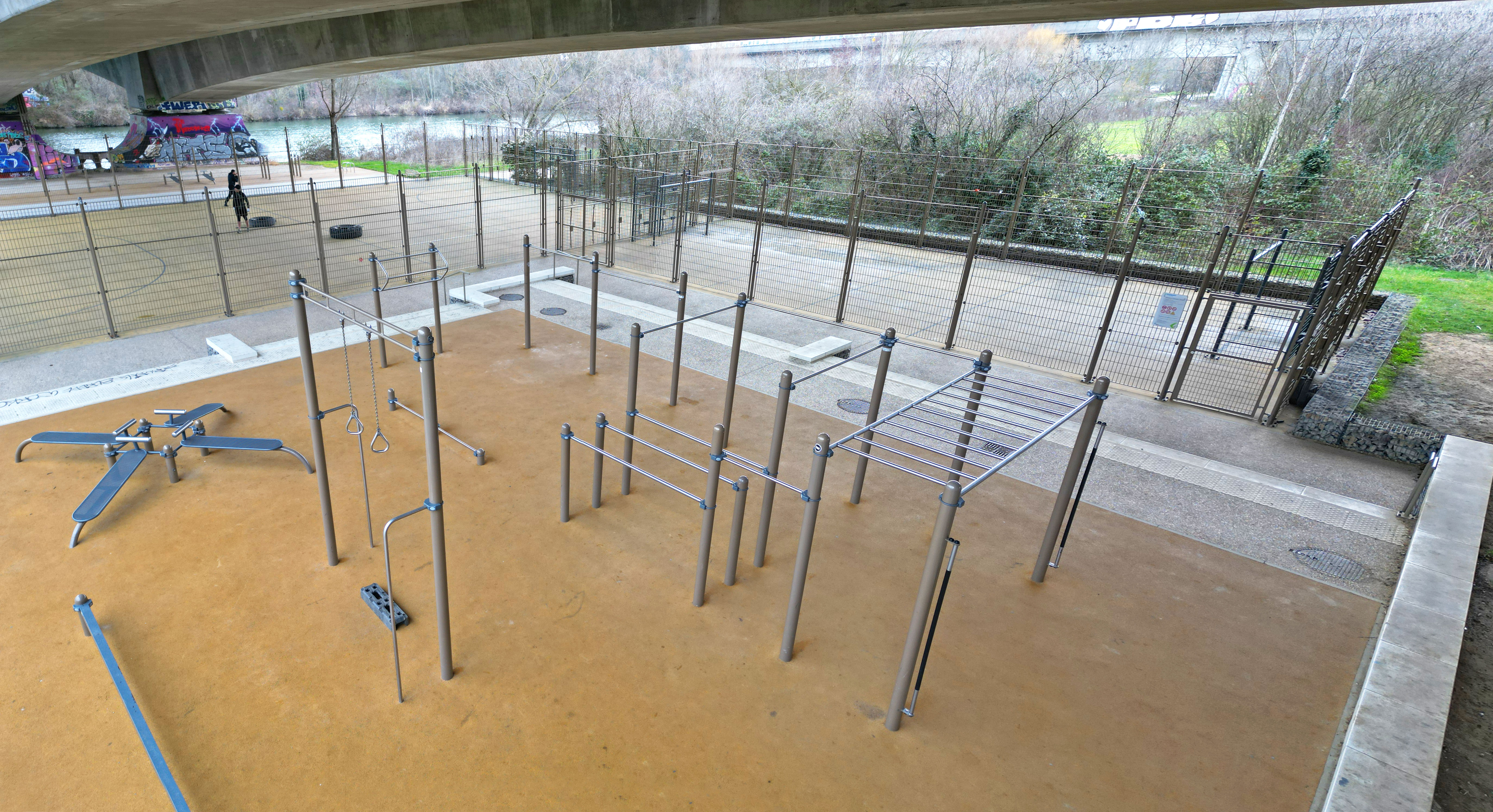 Un parc de fitness extérieur moderne sous un pont avec différents équipements de gymnastique suédoise, dont des structures d'escalade et des barres d'entraînement.