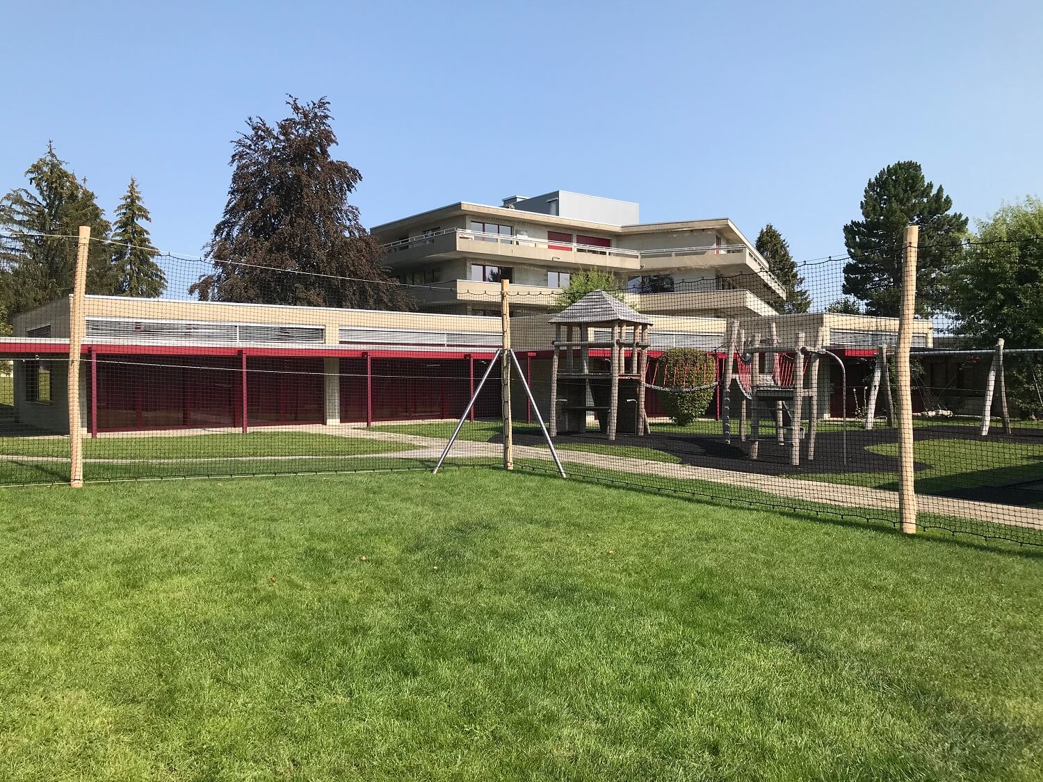 Ein moderner Spielplatz mit Klettergerüst und Sicherheitsnetz auf einem gepflegten Rasen, umgeben von Gebäuden und Bäumen.