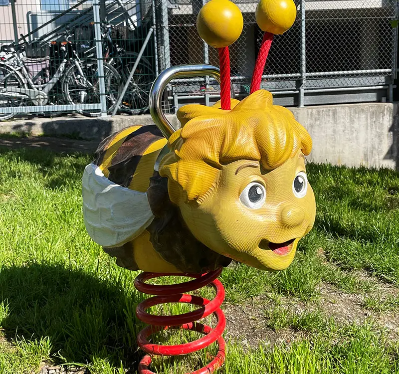 Eine gelbe Wippe auf einem Spielplatz, die wie eine Biene geformt ist, mit einem freundlichen Gesicht und roten Spiralfedern. Hinter der Spielfigur sind Fahrräder und ein Zaun zu sehen.