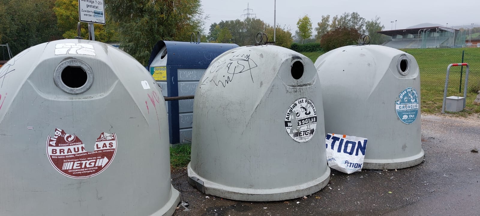Drei große Altglascontainer an einem Sammelplatz im Freien, umgeben von Bäumen und einem Sportplatz im Hintergrund. Sie sind mit Aufklebern beschriftet für Braunglas, Weißglas und Grünglas.